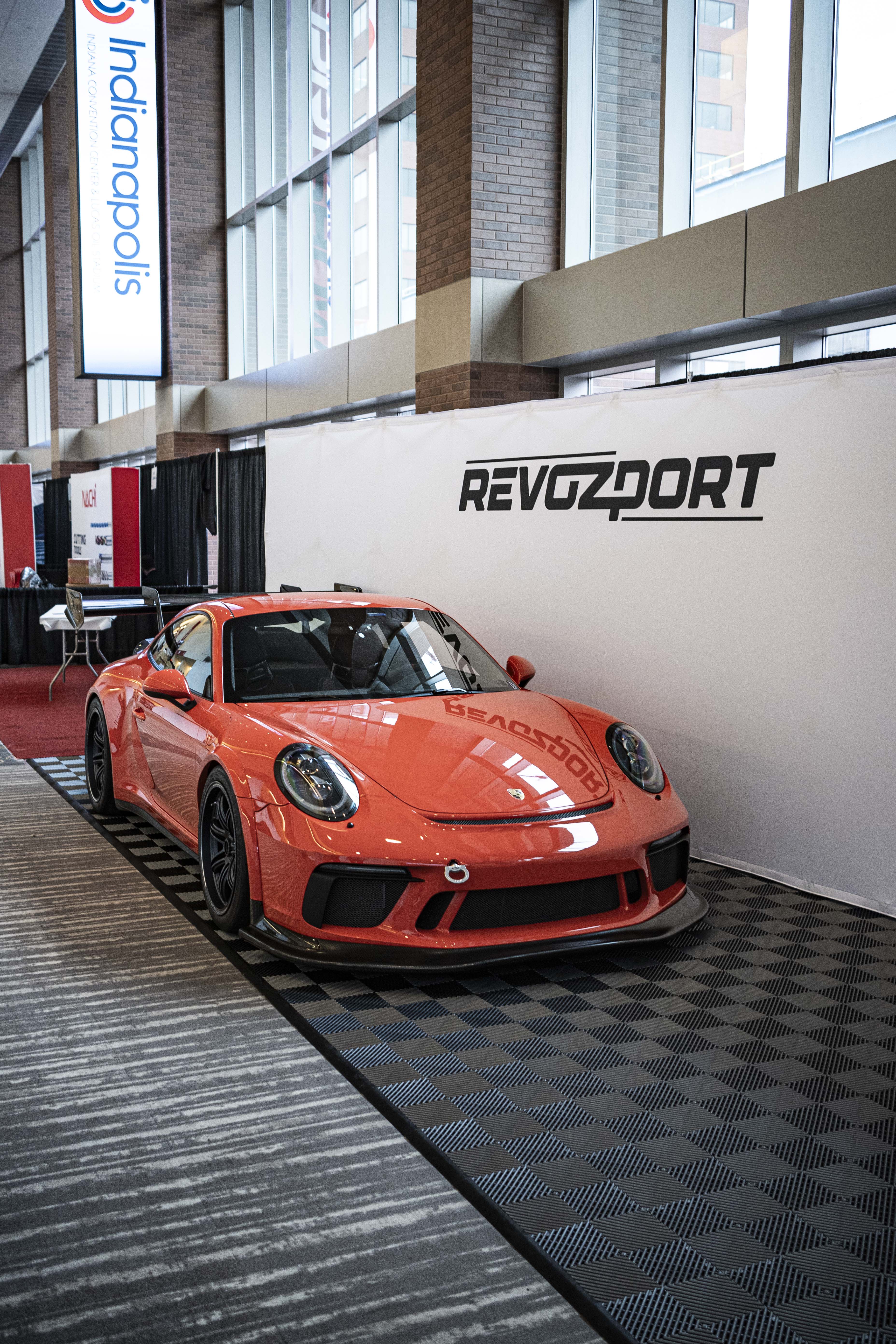 An orange Porsche sports car displayed indoors at an automotive exhibition booth, featuring an aggressive front splitter, black performance wheels, and a sleek aerodynamic body kit. The car is parked in front of a REVOZPORT branded display wall inside a modern convention center with large windows and exhibition setups in the background.