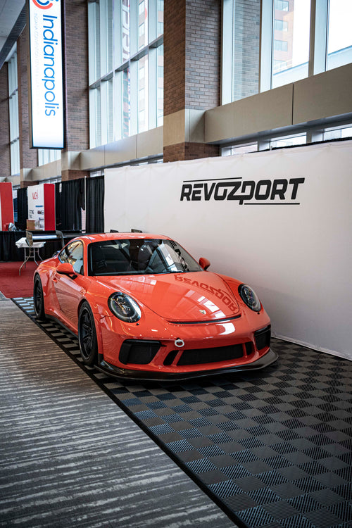 An orange Porsche sports car displayed indoors at an automotive exhibition booth, featuring an aggressive front splitter, black performance wheels, and a sleek aerodynamic body kit. The car is parked in front of a REVOZPORT branded display wall inside a modern convention center with large windows and exhibition setups in the background.