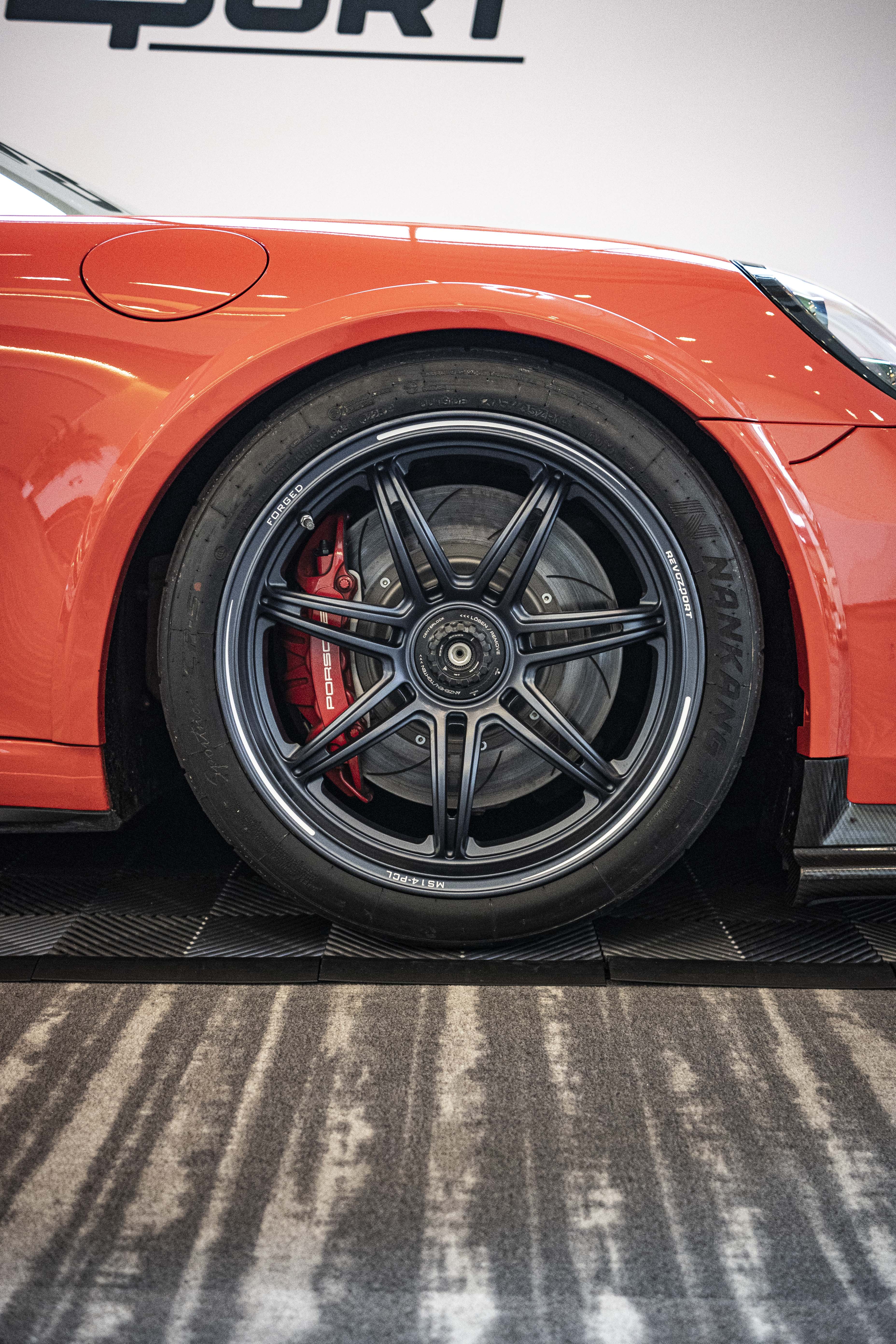 Close-up of a performance wheel setup on an orange Porsche sports car, featuring black multi-spoke forged rims, red brake calipers, and a large drilled brake rotor. The image highlights the aggressive fitment, low-profile tire, and carbon fiber side skirt details at an indoor automotive display.