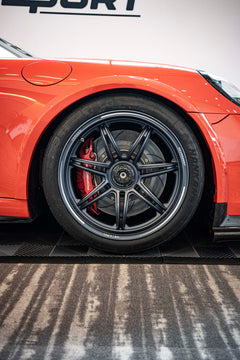 An orange Porsche sports car displayed indoors at an automotive exhibition booth, featuring an aggressive front splitter, black performance wheels, and a sleek aerodynamic body kit. The car is parked in front of a REVOZPORT branded display wall inside a modern convention center with large windows and exhibition setups in the background.