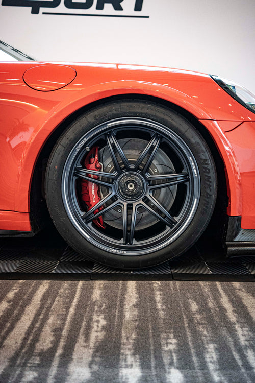 An orange Porsche sports car displayed indoors at an automotive exhibition booth, featuring an aggressive front splitter, black performance wheels, and a sleek aerodynamic body kit. The car is parked in front of a REVOZPORT branded display wall inside a modern convention center with large windows and exhibition setups in the background.