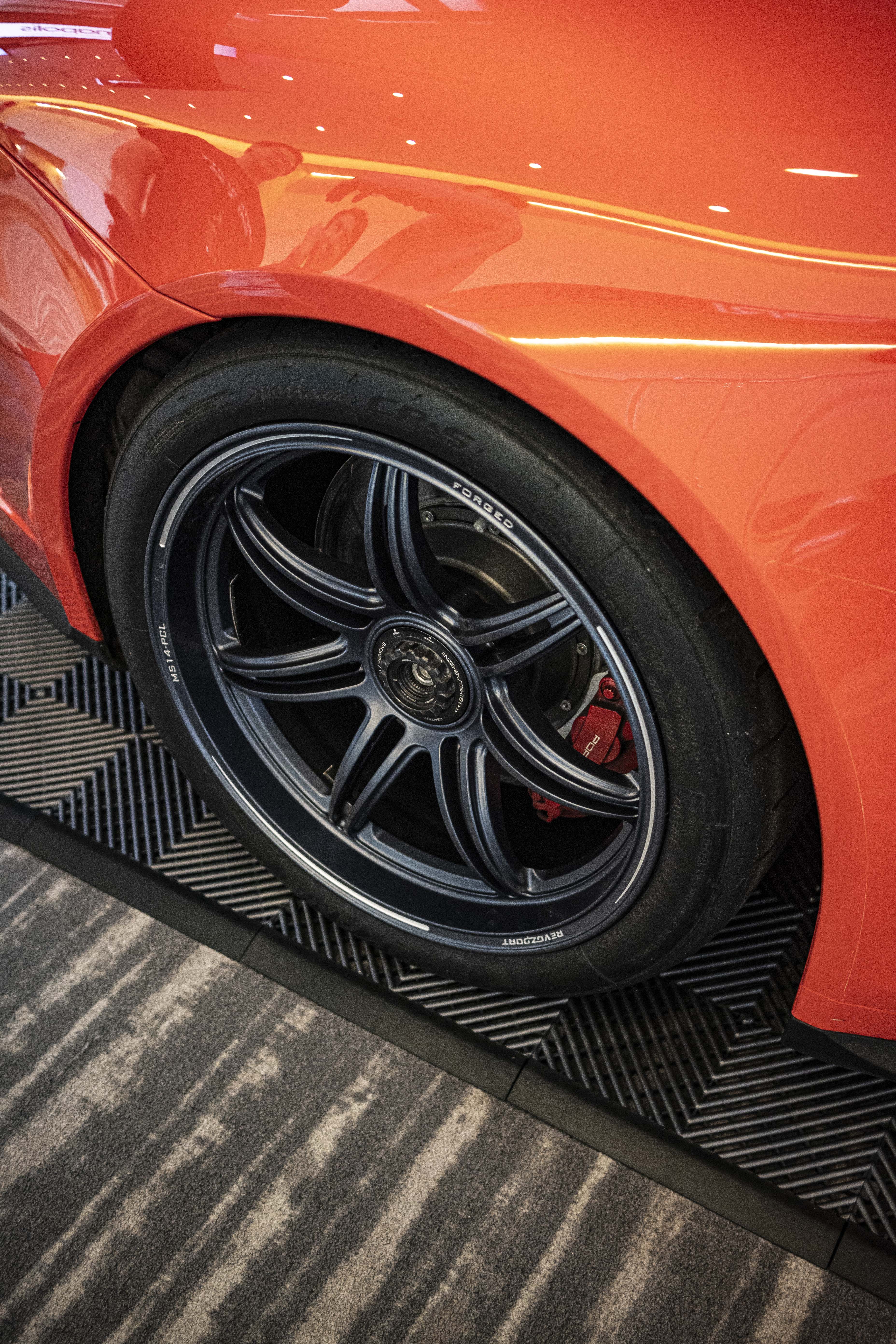 Close-up of a performance wheel setup on an orange Porsche sports car, featuring black multi-spoke forged rims, red brake calipers, and a large drilled brake rotor. The image highlights the aggressive fitment, low-profile tire, and carbon fiber side skirt details at an indoor automotive display.
