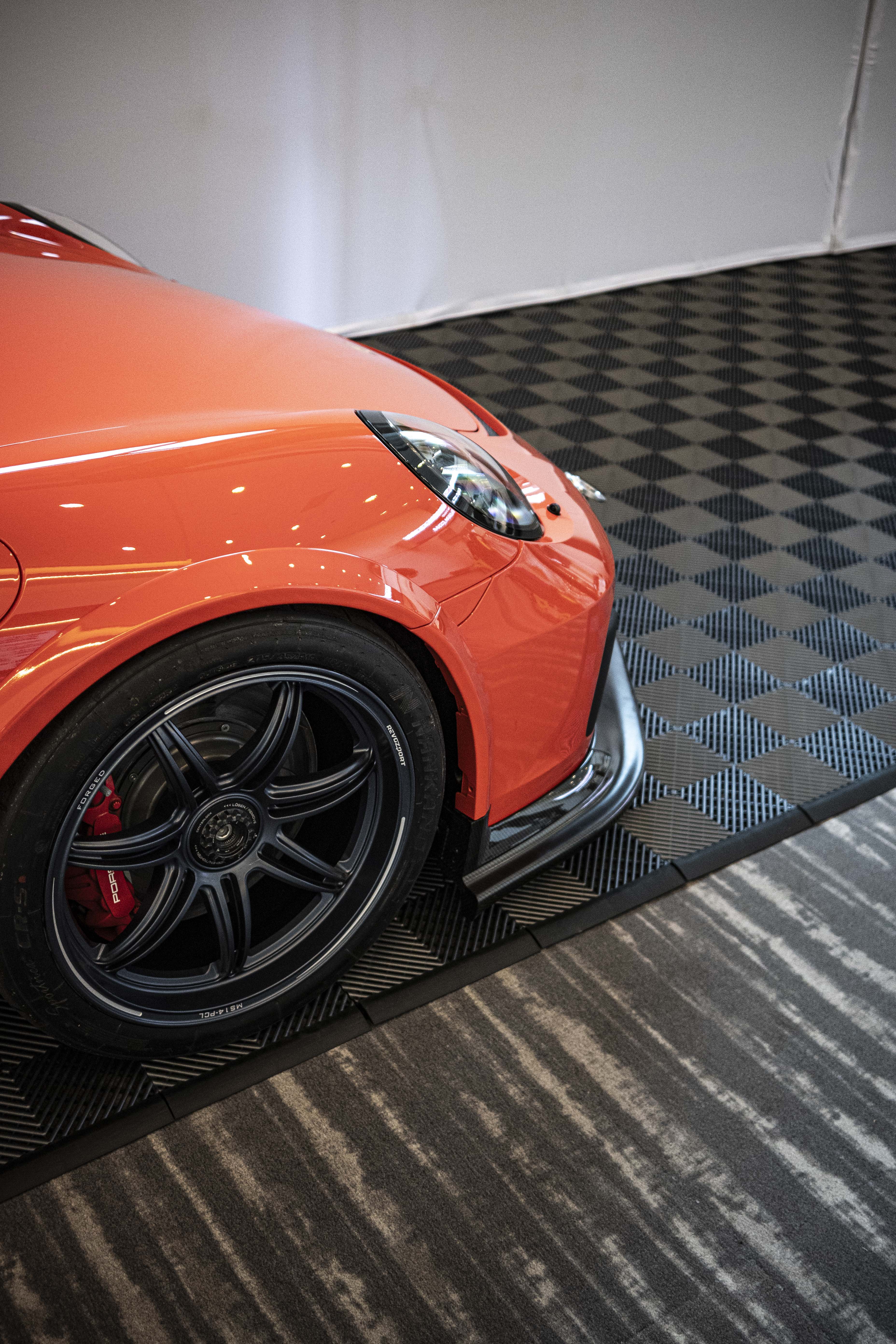 Close-up of a performance wheel setup on an orange Porsche sports car, featuring black multi-spoke forged rims, red brake calipers, and a large drilled brake rotor. The image highlights the aggressive fitment, low-profile tire, and carbon fiber side skirt details at an indoor automotive display.