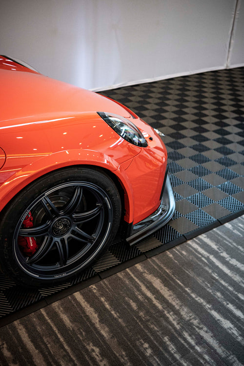 Close-up of a performance wheel setup on an orange Porsche sports car, featuring black multi-spoke forged rims, red brake calipers, and a large drilled brake rotor. The image highlights the aggressive fitment, low-profile tire, and carbon fiber side skirt details at an indoor automotive display.