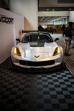 Silver Chevrolet Corvette race car displayed at an indoor automotive exhibition, featuring a wide-body aerodynamic kit, large rear wing, carbon fiber front splitter, and vented hood. The aggressive motorsport styling is showcased under exhibition lighting with REVOZPORT branding in the background.