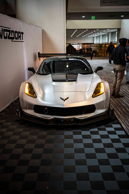 Silver Chevrolet Corvette race car displayed at an indoor automotive exhibition, featuring a wide-body aerodynamic kit, large rear wing, carbon fiber front splitter, and vented hood. The aggressive motorsport styling is showcased under exhibition lighting with REVOZPORT branding in the background.