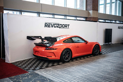 An orange Porsche sports car displayed indoors at an automotive exhibition booth, featuring an aggressive front splitter, black performance wheels, and a sleek aerodynamic body kit. The car is parked in front of a REVOZPORT branded display wall inside a modern convention center with large windows and exhibition setups in the background.