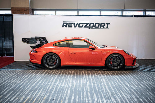 An orange Porsche sports car displayed indoors at an automotive exhibition booth, featuring an aggressive front splitter, black performance wheels, and a sleek aerodynamic body kit. The car is parked in front of a REVOZPORT branded display wall inside a modern convention center with large windows and exhibition setups in the background.