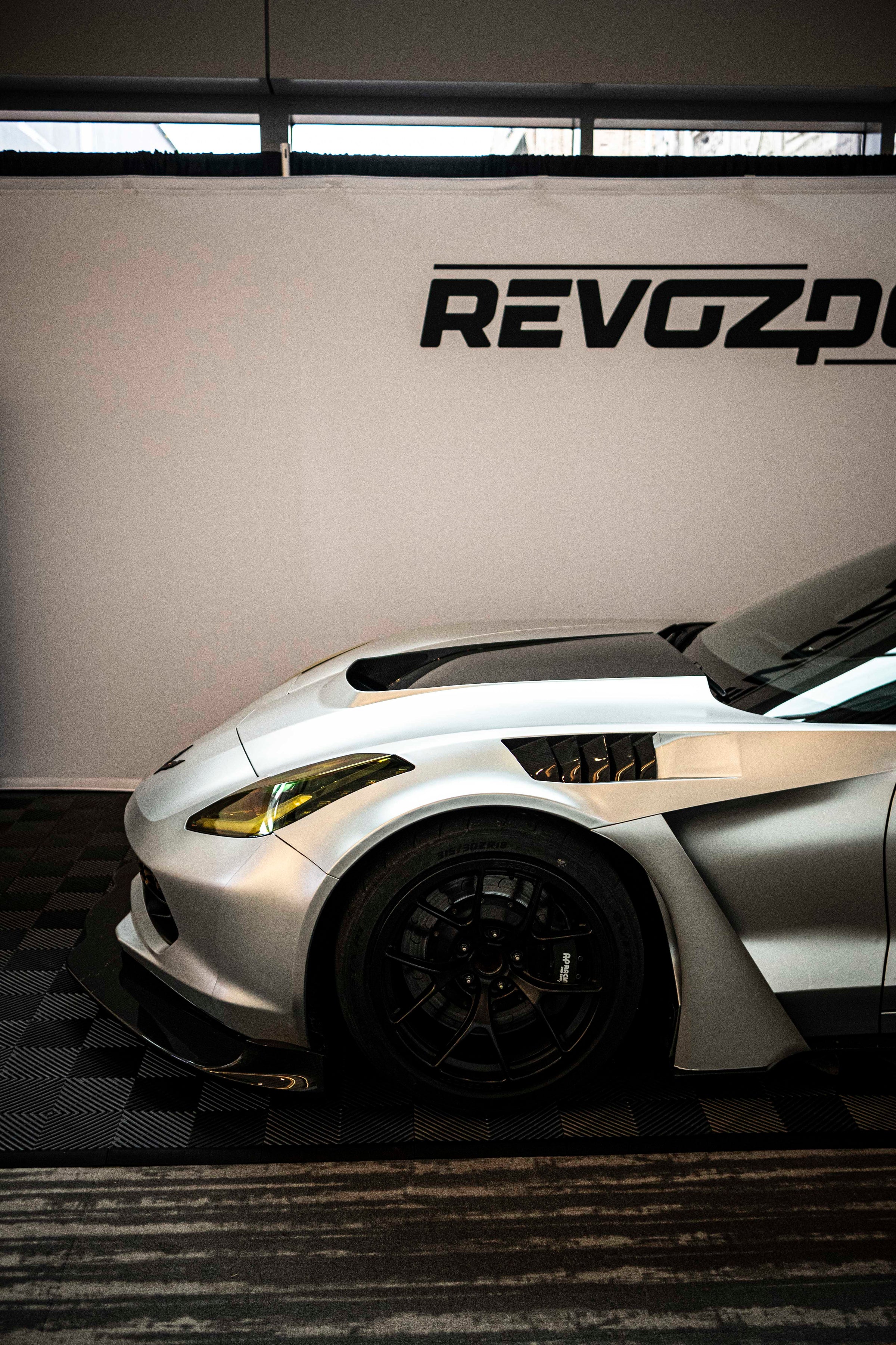 Silver Chevrolet Corvette race car displayed at an indoor automotive exhibition, featuring a wide-body aerodynamic kit, large rear wing, carbon fiber front splitter, and vented hood. The aggressive motorsport styling is showcased under exhibition lighting with REVOZPORT branding in the background.