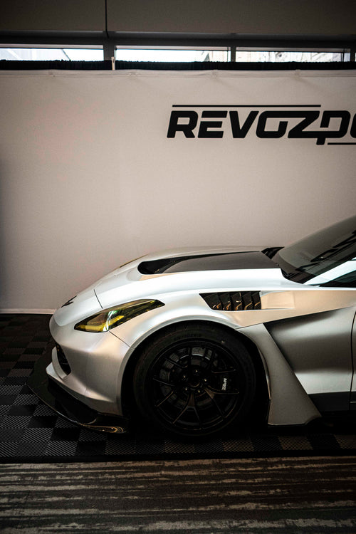 Silver Chevrolet Corvette race car displayed at an indoor automotive exhibition, featuring a wide-body aerodynamic kit, large rear wing, carbon fiber front splitter, and vented hood. The aggressive motorsport styling is showcased under exhibition lighting with REVOZPORT branding in the background.