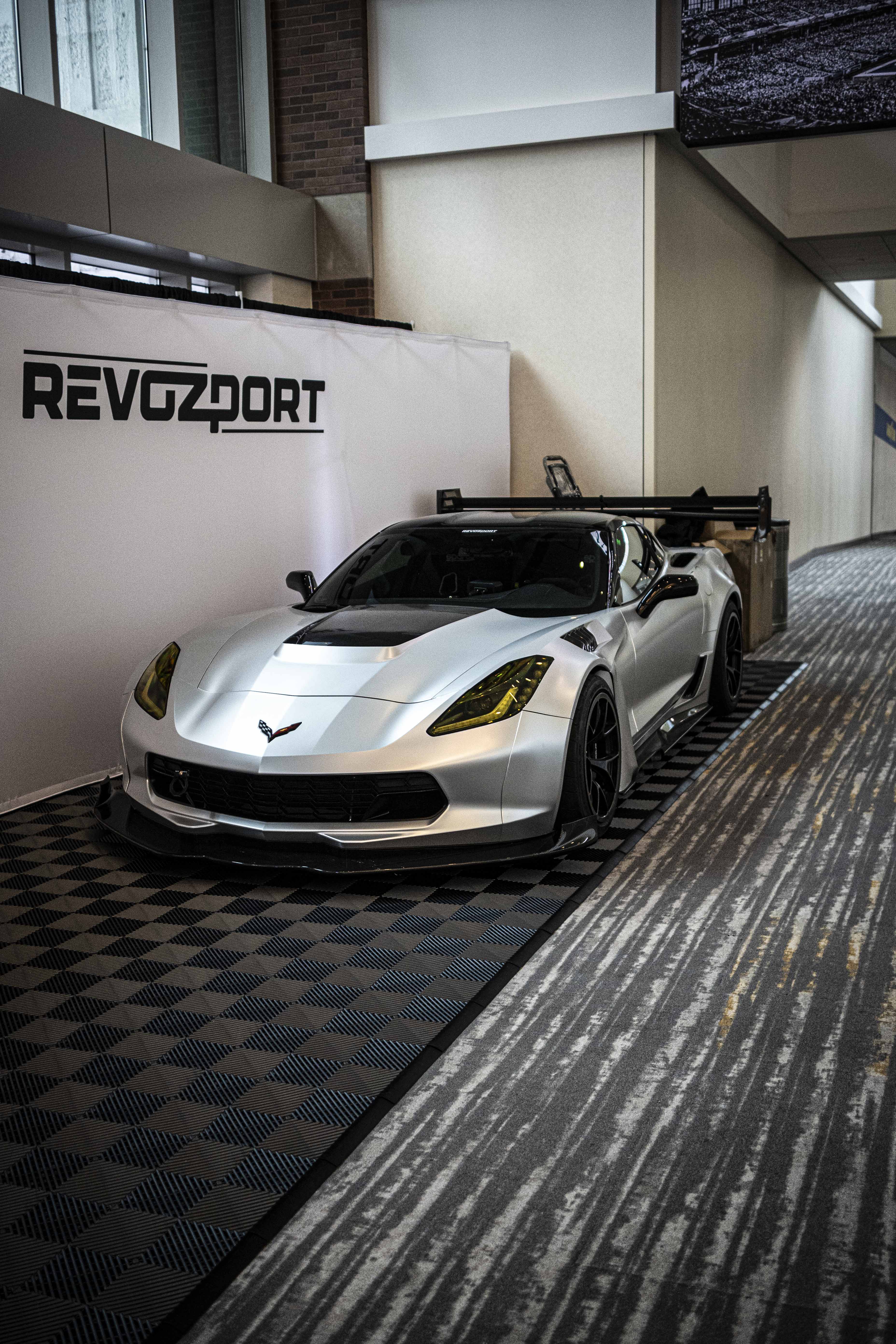 Silver Chevrolet Corvette race car displayed at an indoor automotive exhibition, featuring a wide-body aerodynamic kit, large rear wing, carbon fiber front splitter, and vented hood. The aggressive motorsport styling is showcased under exhibition lighting with REVOZPORT branding in the background.