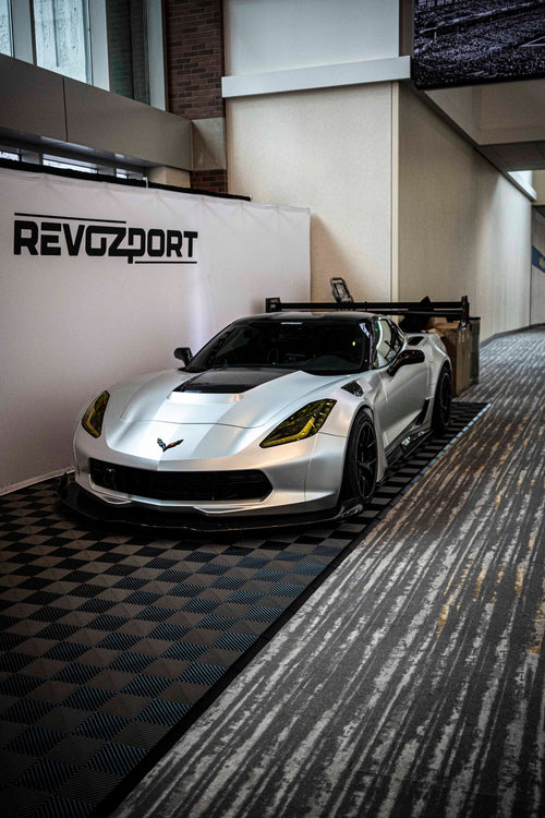 Silver Chevrolet Corvette race car displayed at an indoor automotive exhibition, featuring a wide-body aerodynamic kit, large rear wing, carbon fiber front splitter, and vented hood. The aggressive motorsport styling is showcased under exhibition lighting with REVOZPORT branding in the background.