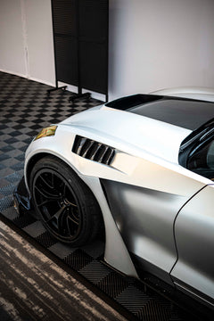 Silver Chevrolet Corvette race car displayed at an indoor automotive exhibition, featuring a wide-body aerodynamic kit, large rear wing, carbon fiber front splitter, and vented hood. The aggressive motorsport styling is showcased under exhibition lighting with REVOZPORT branding in the background.