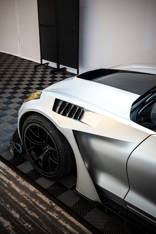 Silver Chevrolet Corvette race car displayed at an indoor automotive exhibition, featuring a wide-body aerodynamic kit, large rear wing, carbon fiber front splitter, and vented hood. The aggressive motorsport styling is showcased under exhibition lighting with REVOZPORT branding in the background.