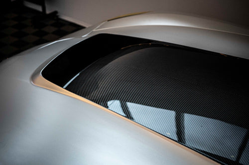 Silver Chevrolet Corvette race car displayed at an indoor automotive exhibition, featuring a wide-body aerodynamic kit, large rear wing, carbon fiber front splitter, and vented hood. The aggressive motorsport styling is showcased under exhibition lighting with REVOZPORT branding in the background.