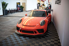 An orange Porsche sports car displayed indoors at an automotive exhibition booth, featuring an aggressive front splitter, black performance wheels, and a sleek aerodynamic body kit. The car is parked in front of a REVOZPORT branded display wall inside a modern convention center with large windows and exhibition setups in the background.