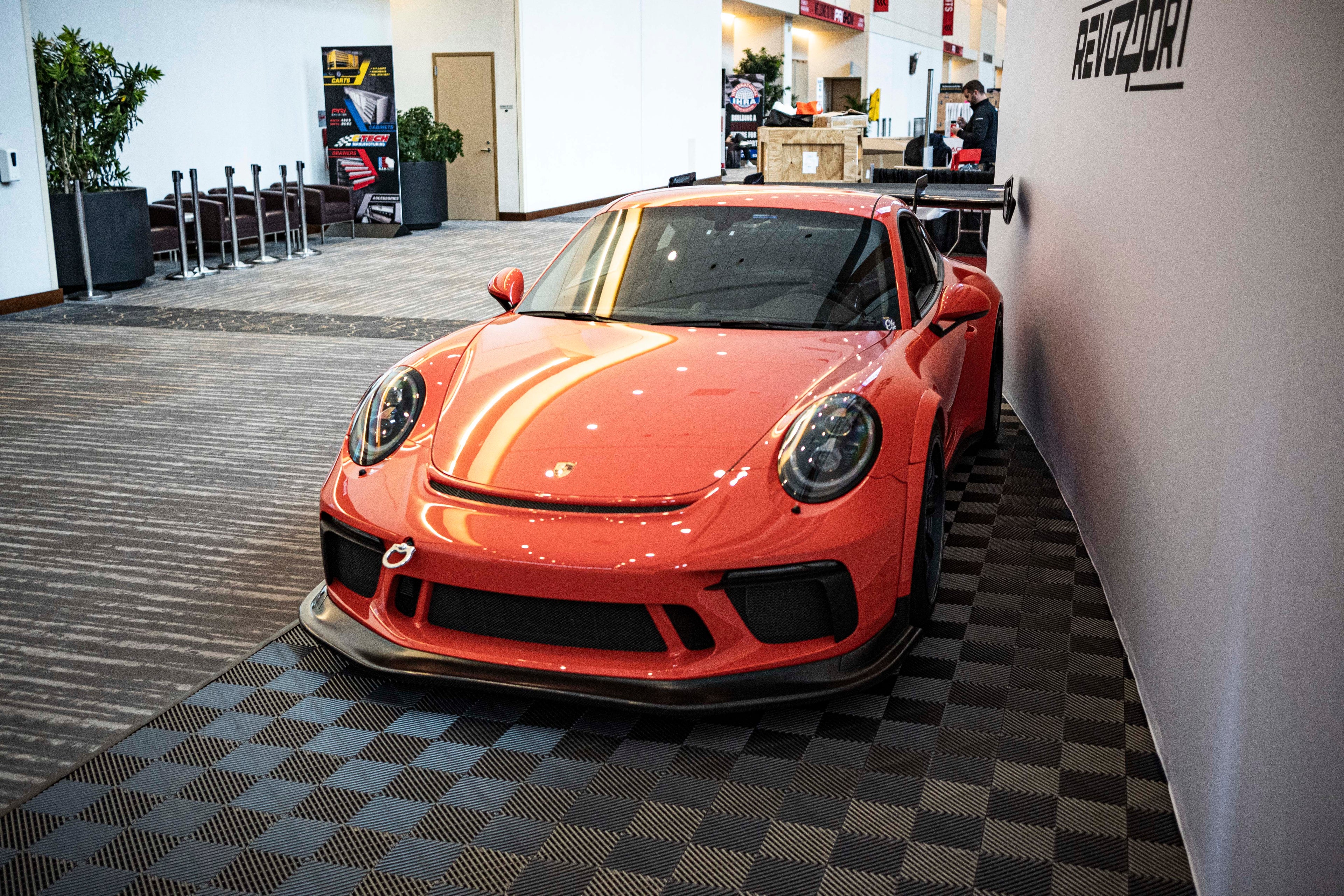 An orange Porsche sports car displayed indoors at an automotive exhibition booth, featuring an aggressive front splitter, black performance wheels, and a sleek aerodynamic body kit. The car is parked in front of a REVOZPORT branded display wall inside a modern convention center with large windows and exhibition setups in the background.