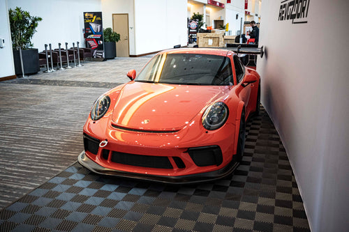 An orange Porsche sports car displayed indoors at an automotive exhibition booth, featuring an aggressive front splitter, black performance wheels, and a sleek aerodynamic body kit. The car is parked in front of a REVOZPORT branded display wall inside a modern convention center with large windows and exhibition setups in the background.