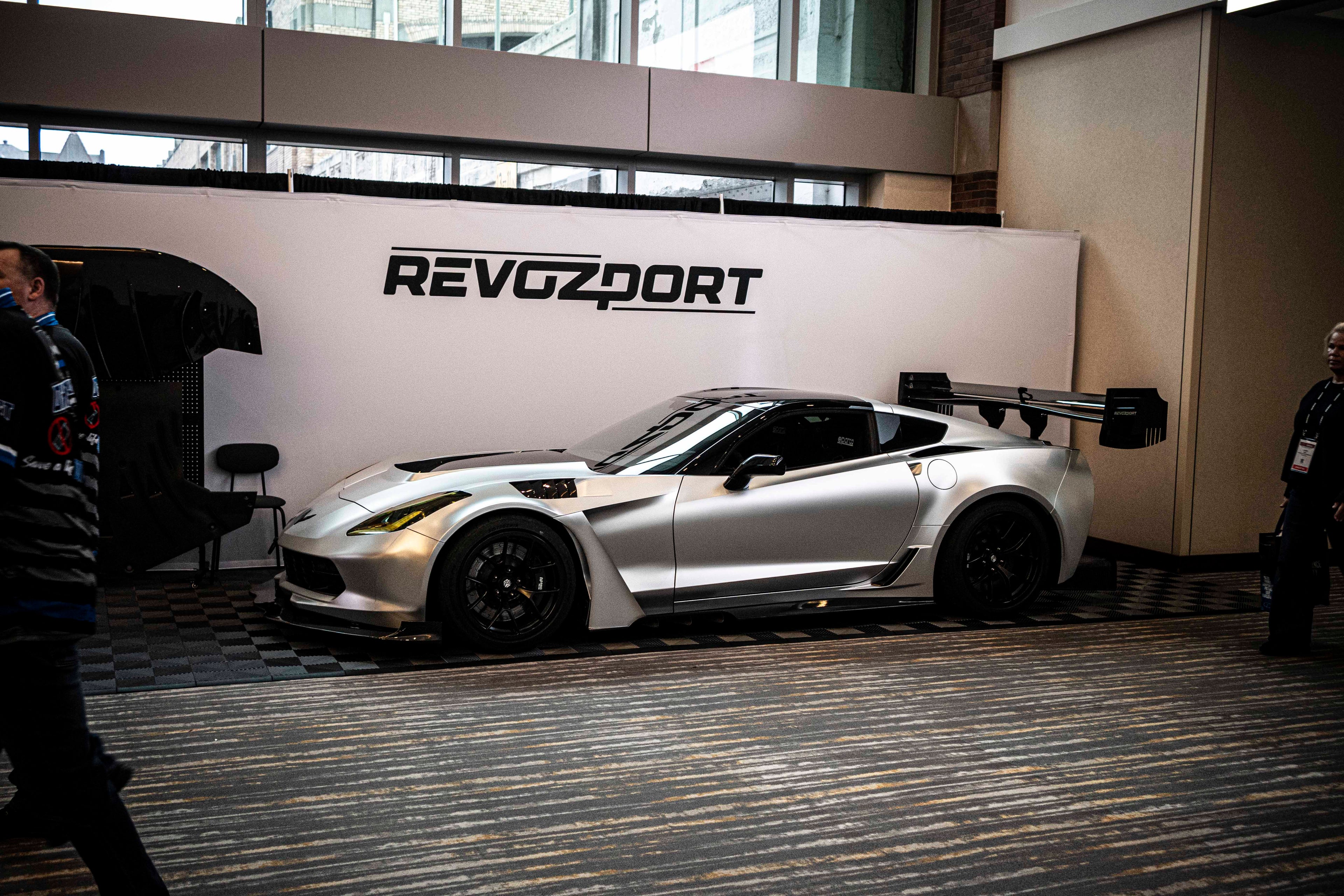 Silver Chevrolet Corvette race car displayed at an indoor automotive exhibition, featuring a wide-body aerodynamic kit, large rear wing, carbon fiber front splitter, and vented hood. The aggressive motorsport styling is showcased under exhibition lighting with REVOZPORT branding in the background.