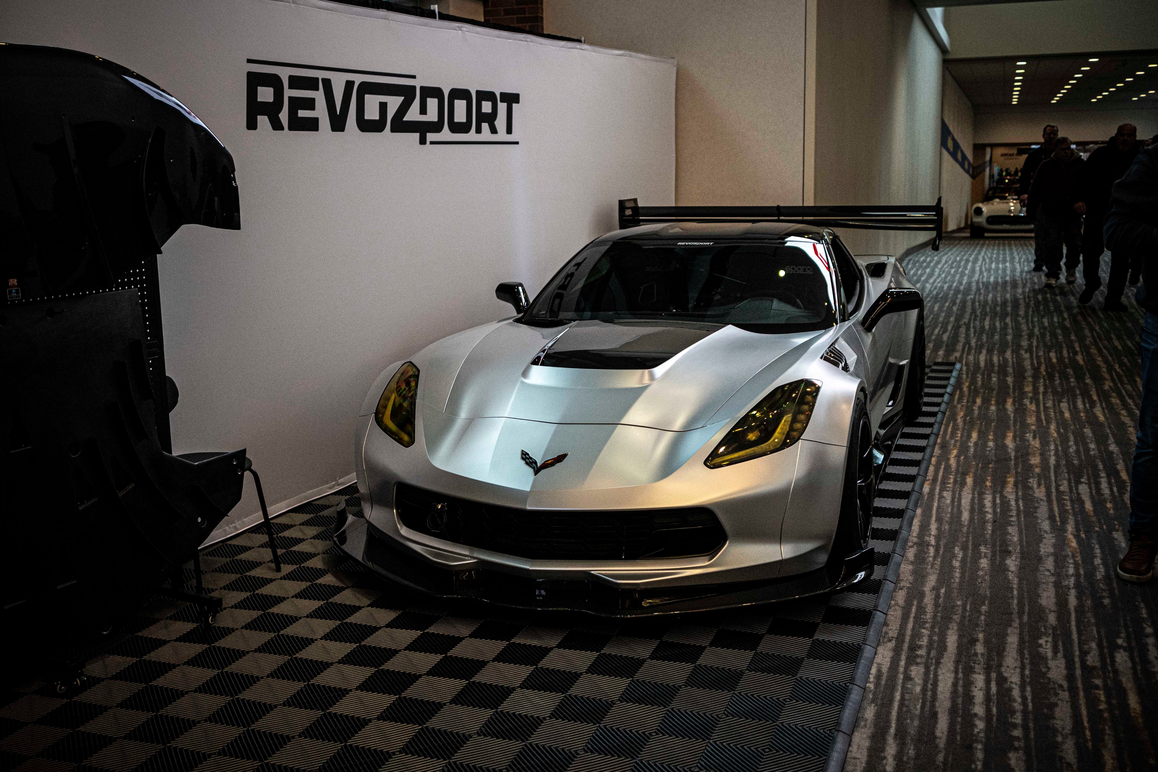 Silver Chevrolet Corvette race car displayed at an indoor automotive exhibition, featuring a wide-body aerodynamic kit, large rear wing, carbon fiber front splitter, and vented hood. The aggressive motorsport styling is showcased under exhibition lighting with REVOZPORT branding in the background.