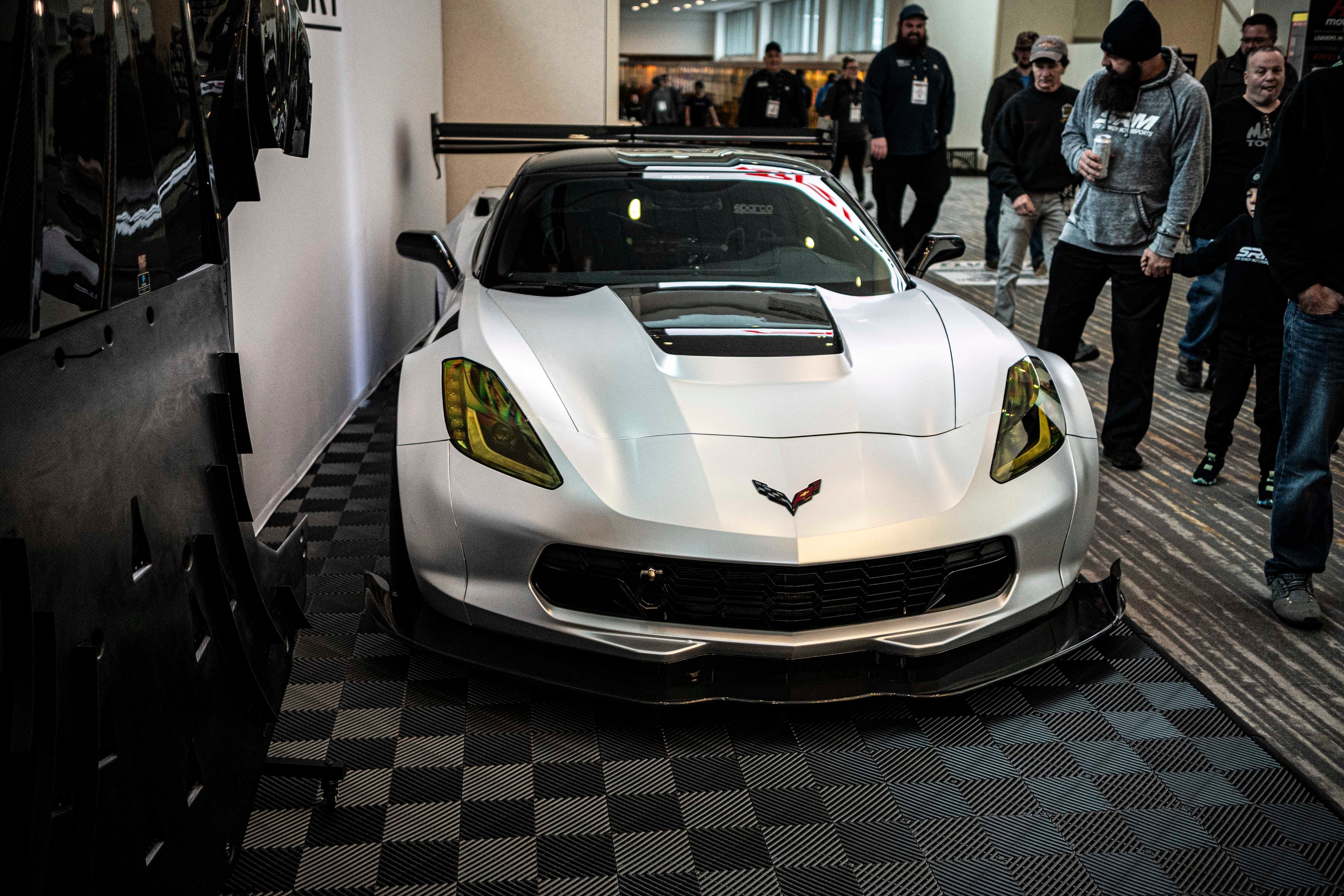 Silver Chevrolet Corvette race car displayed at an indoor automotive exhibition, featuring a wide-body aerodynamic kit, large rear wing, carbon fiber front splitter, and vented hood. The aggressive motorsport styling is showcased under exhibition lighting with REVOZPORT branding in the background.