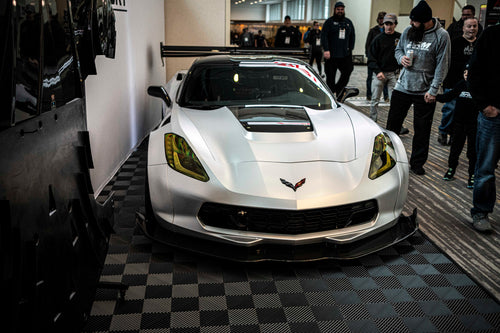 Silver Chevrolet Corvette race car displayed at an indoor automotive exhibition, featuring a wide-body aerodynamic kit, large rear wing, carbon fiber front splitter, and vented hood. The aggressive motorsport styling is showcased under exhibition lighting with REVOZPORT branding in the background.