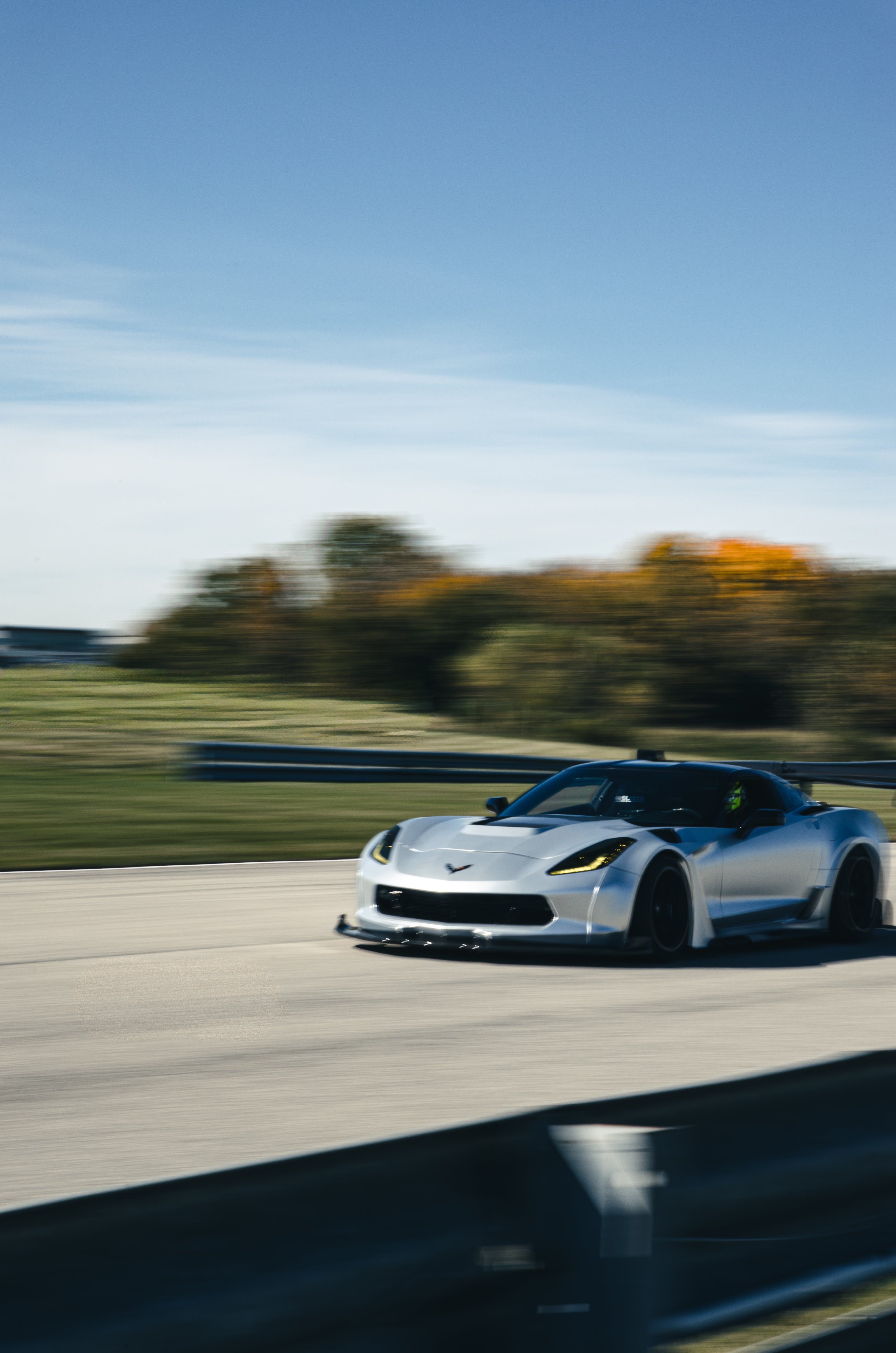 Silver Chevrolet Corvette C7 with carbon fiber front splitter driving on a sunny road, surrounded by autumn trees and a clear blue sky.