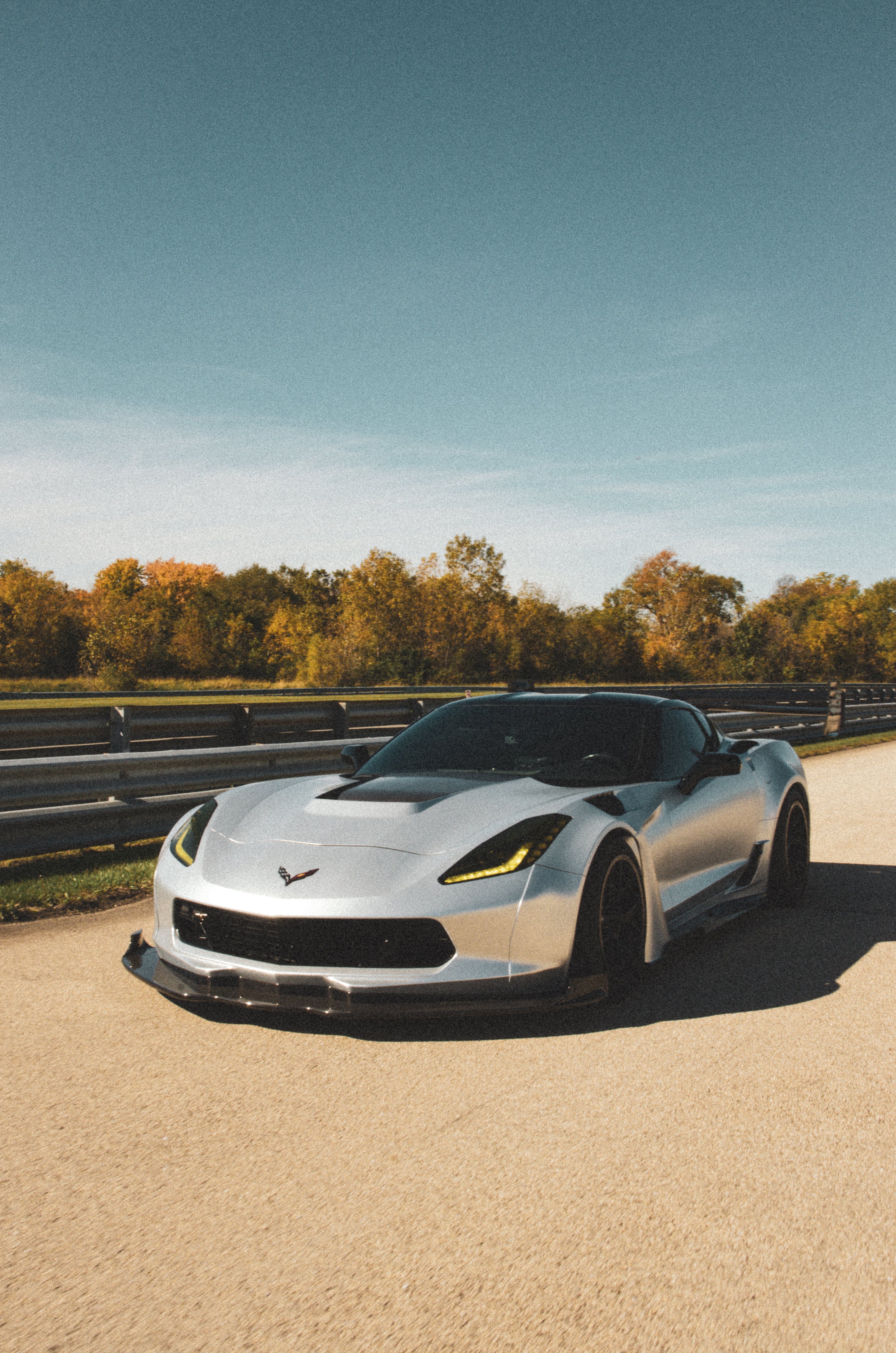 Silver Chevrolet Corvette C7 with carbon fiber front splitter driving on a sunny road, surrounded by autumn trees and a clear blue sky.