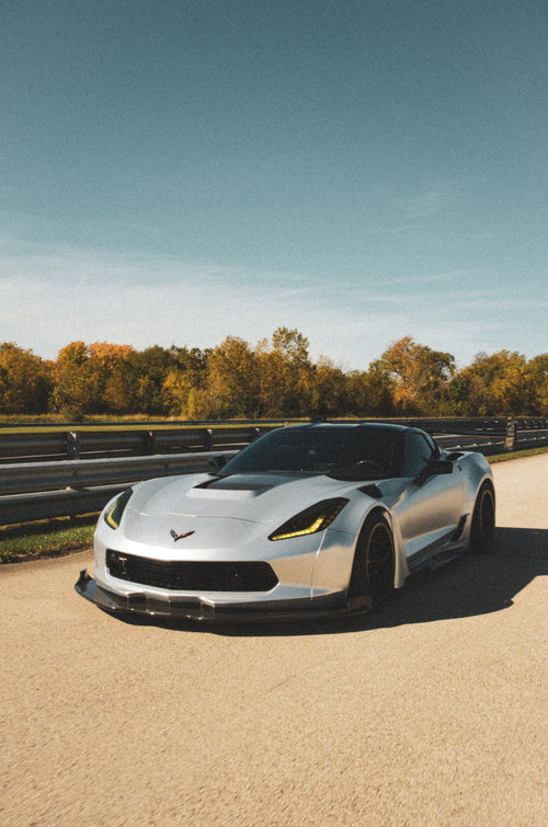 Silver Chevrolet Corvette C7 with carbon fiber front splitter driving on a sunny road, surrounded by autumn trees and a clear blue sky.