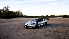 Silver Chevrolet Corvette C7 with carbon fiber front splitter driving on a sunny road, surrounded by autumn trees and a clear blue sky.