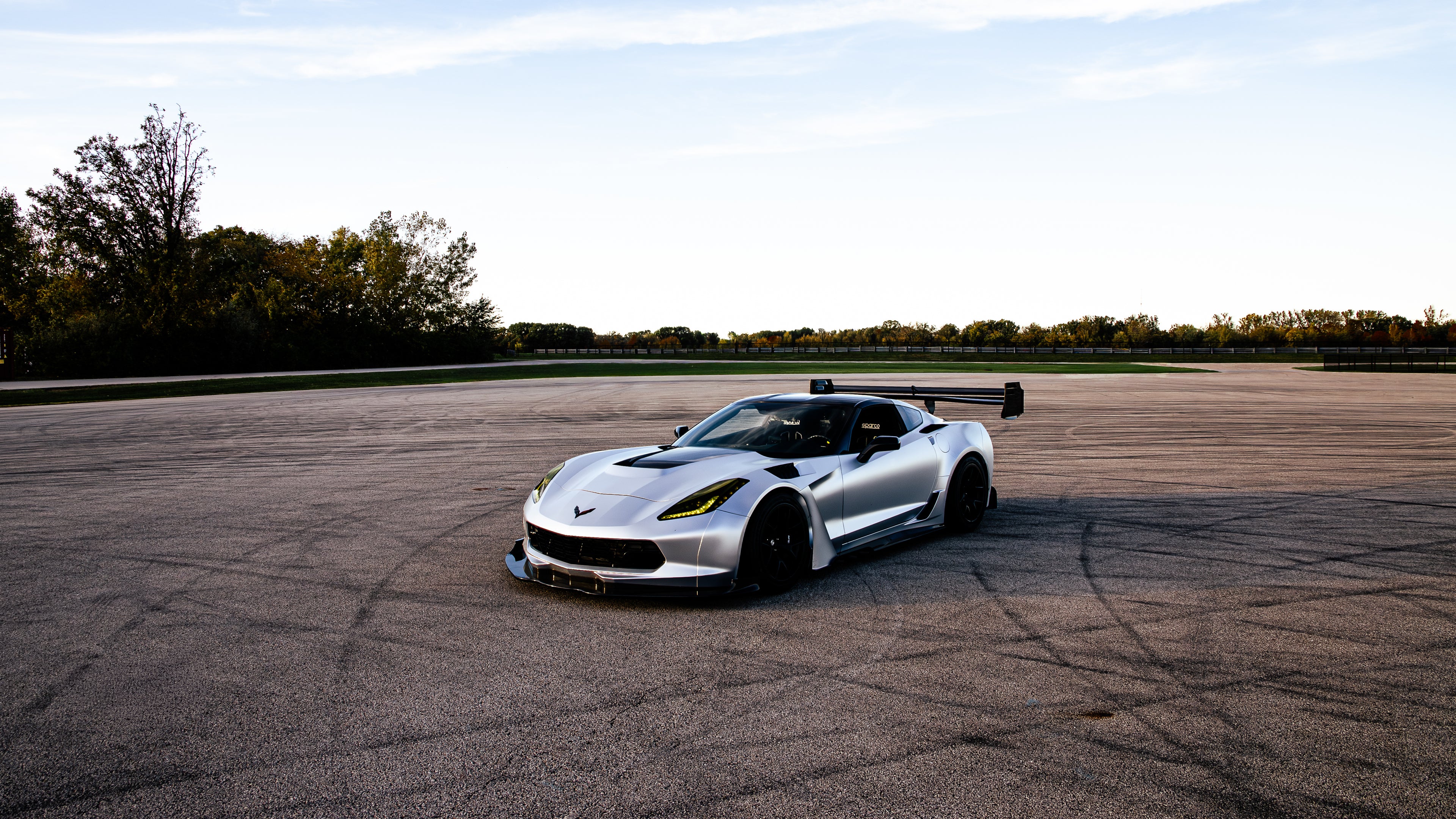 Silver Chevrolet Corvette C7 with carbon fiber front splitter driving on a sunny road, surrounded by autumn trees and a clear blue sky.