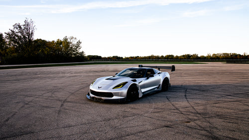 Silver Chevrolet Corvette C7 with carbon fiber front splitter driving on a sunny road, surrounded by autumn trees and a clear blue sky.