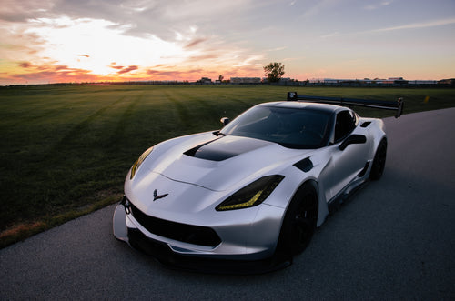 Silver Chevrolet Corvette C7 with carbon fiber front splitter driving on a sunny road, surrounded by autumn trees and a clear blue sky.