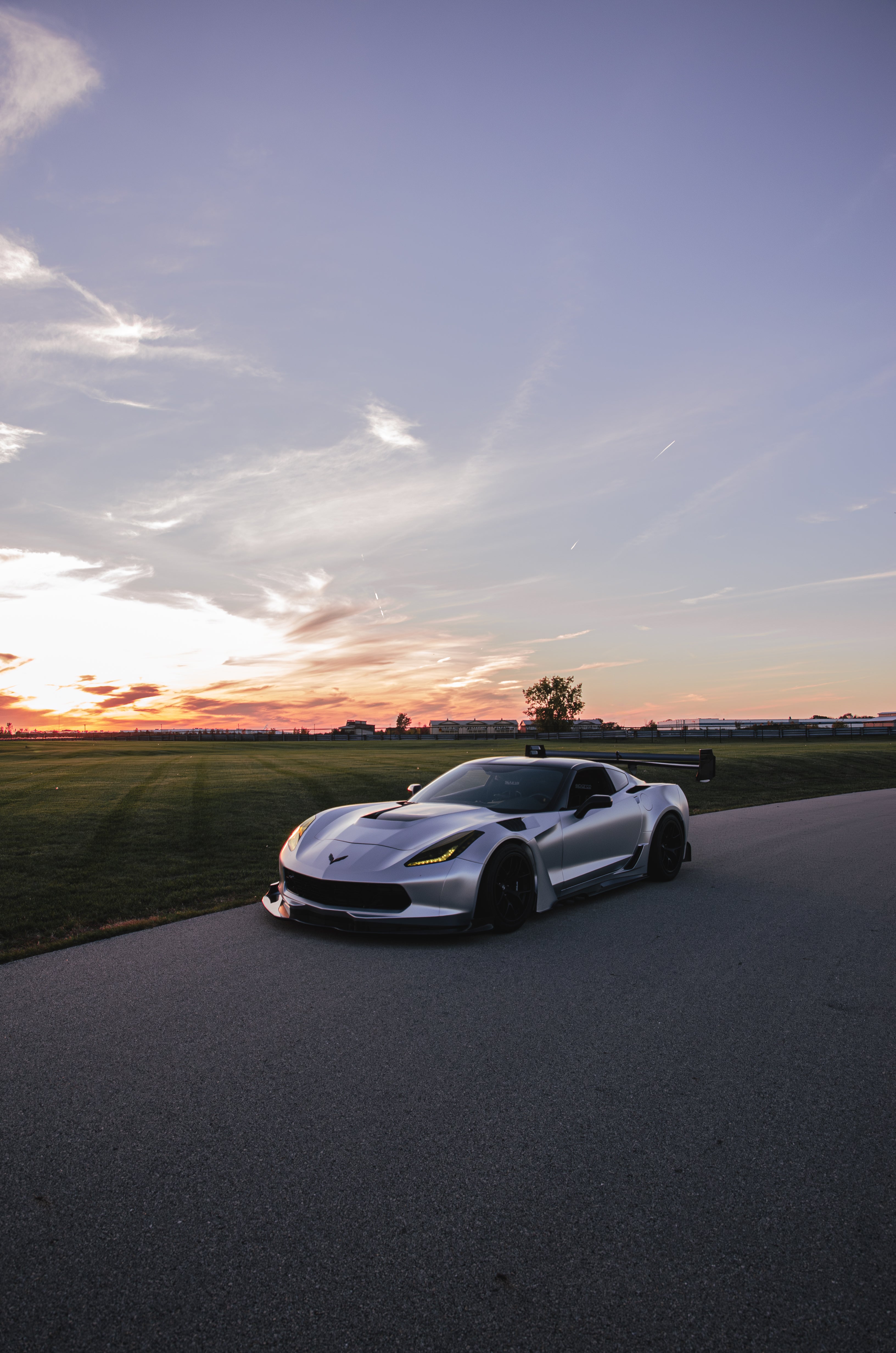 Silver Chevrolet Corvette C7 with carbon fiber front splitter driving on a sunny road, surrounded by autumn trees and a clear blue sky.