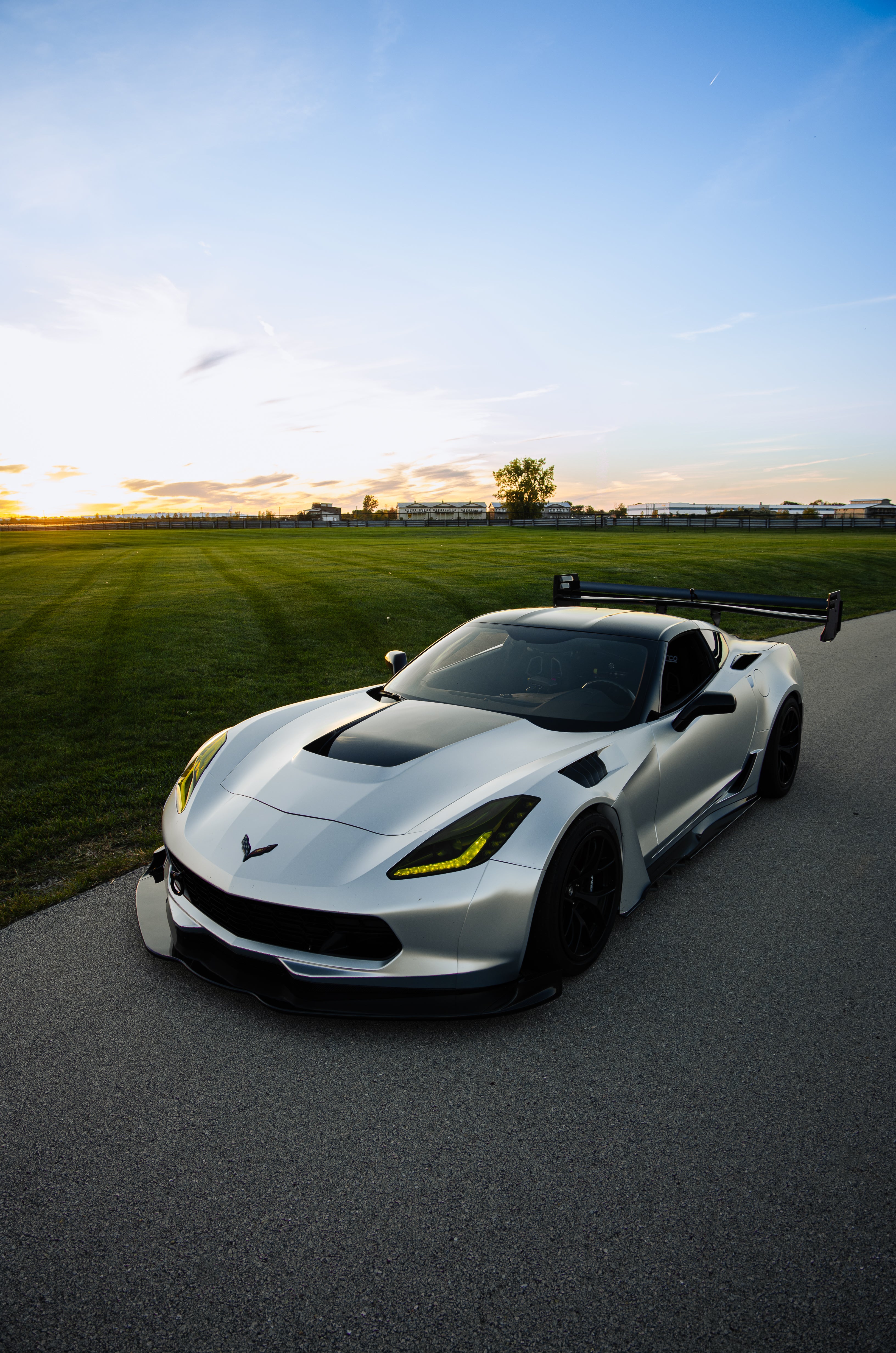 Silver Chevrolet Corvette C7 with carbon fiber front splitter driving on a sunny road, surrounded by autumn trees and a clear blue sky.