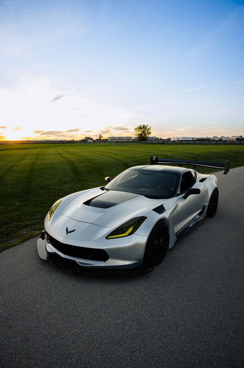 Silver Chevrolet Corvette C7 with carbon fiber front splitter driving on a sunny road, surrounded by autumn trees and a clear blue sky.