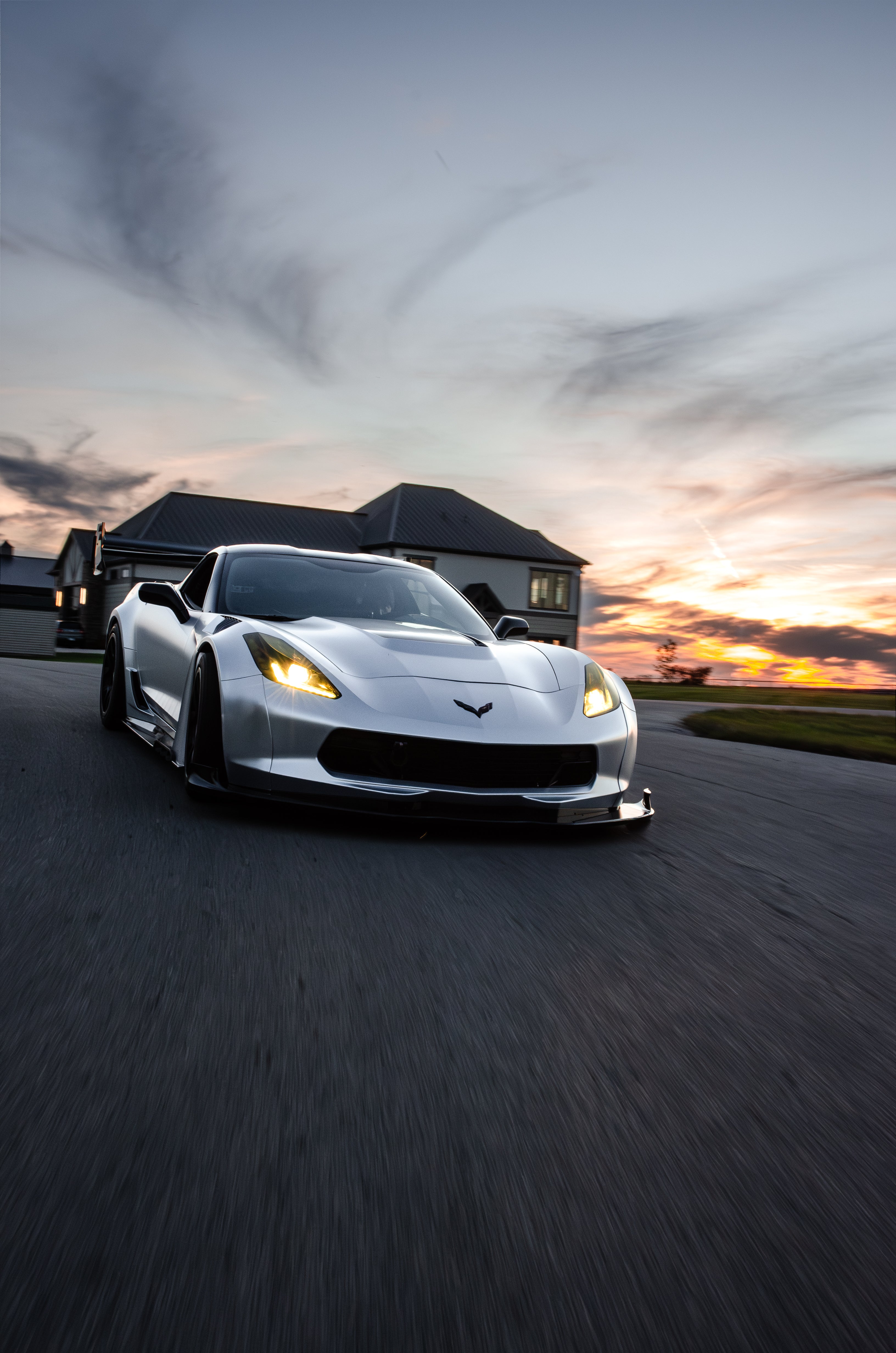 Silver Chevrolet Corvette C7 with carbon fiber front splitter driving on a sunny road, surrounded by autumn trees and a clear blue sky.