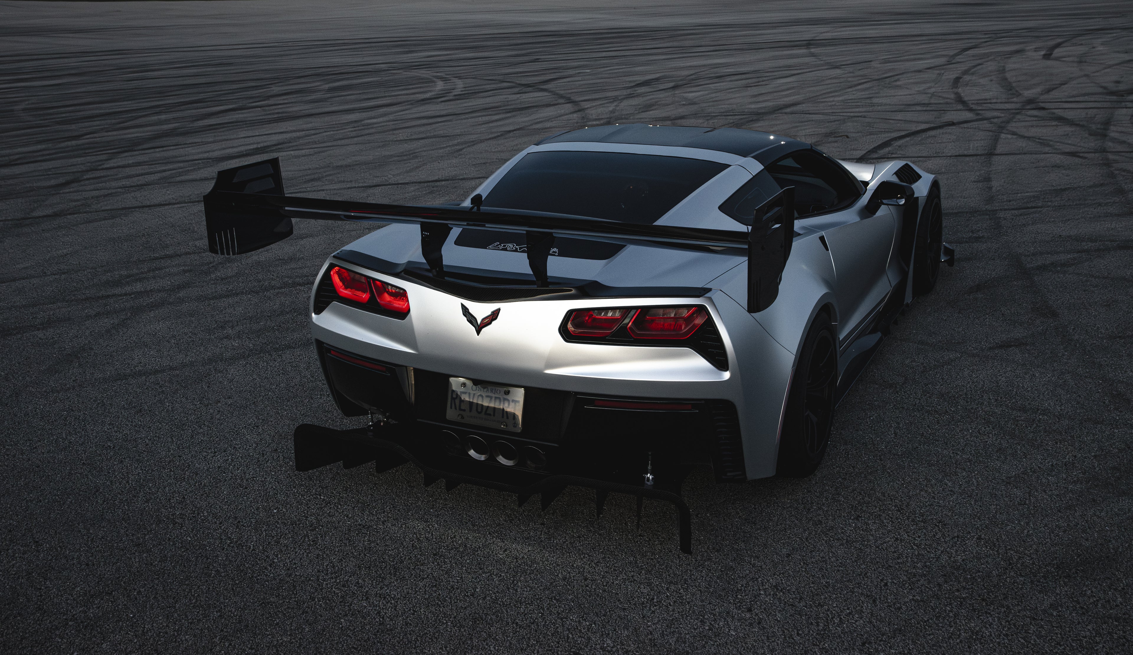 Silver Chevrolet Corvette C7 with carbon fiber front splitter driving on a sunny road, surrounded by autumn trees and a clear blue sky.
