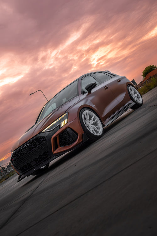Bronze Audi RS3 Sportback featuring a REVOZPORT carbon fiber body kit with front lip and side skirts, paired with white multi-spoke forged wheels, parked at sunset in an open urban setting.