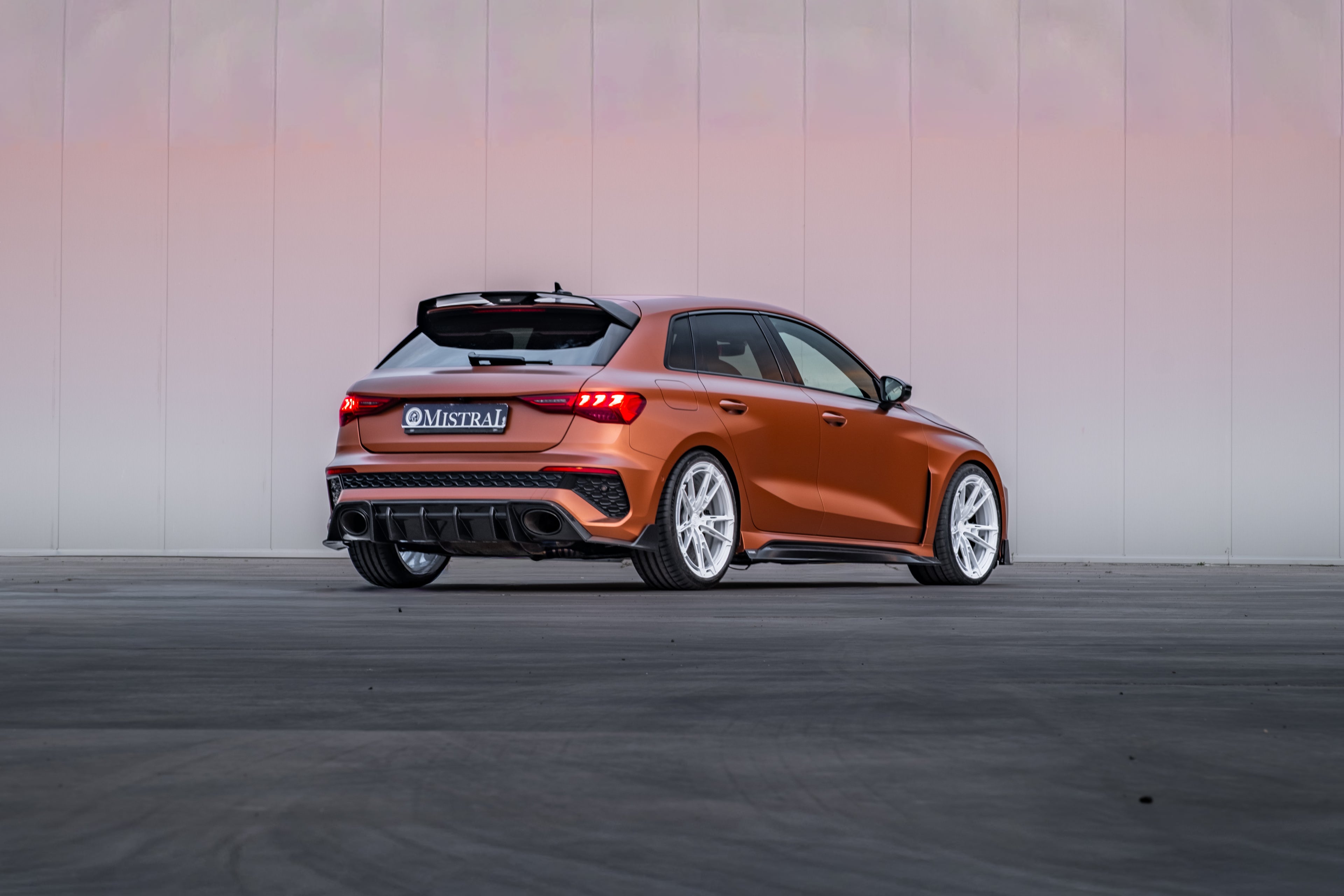 Bronze Audi RS3 Sportback featuring a REVOZPORT carbon fiber body kit with front lip and side skirts, paired with white multi-spoke forged wheels, parked at sunset in an open urban setting.