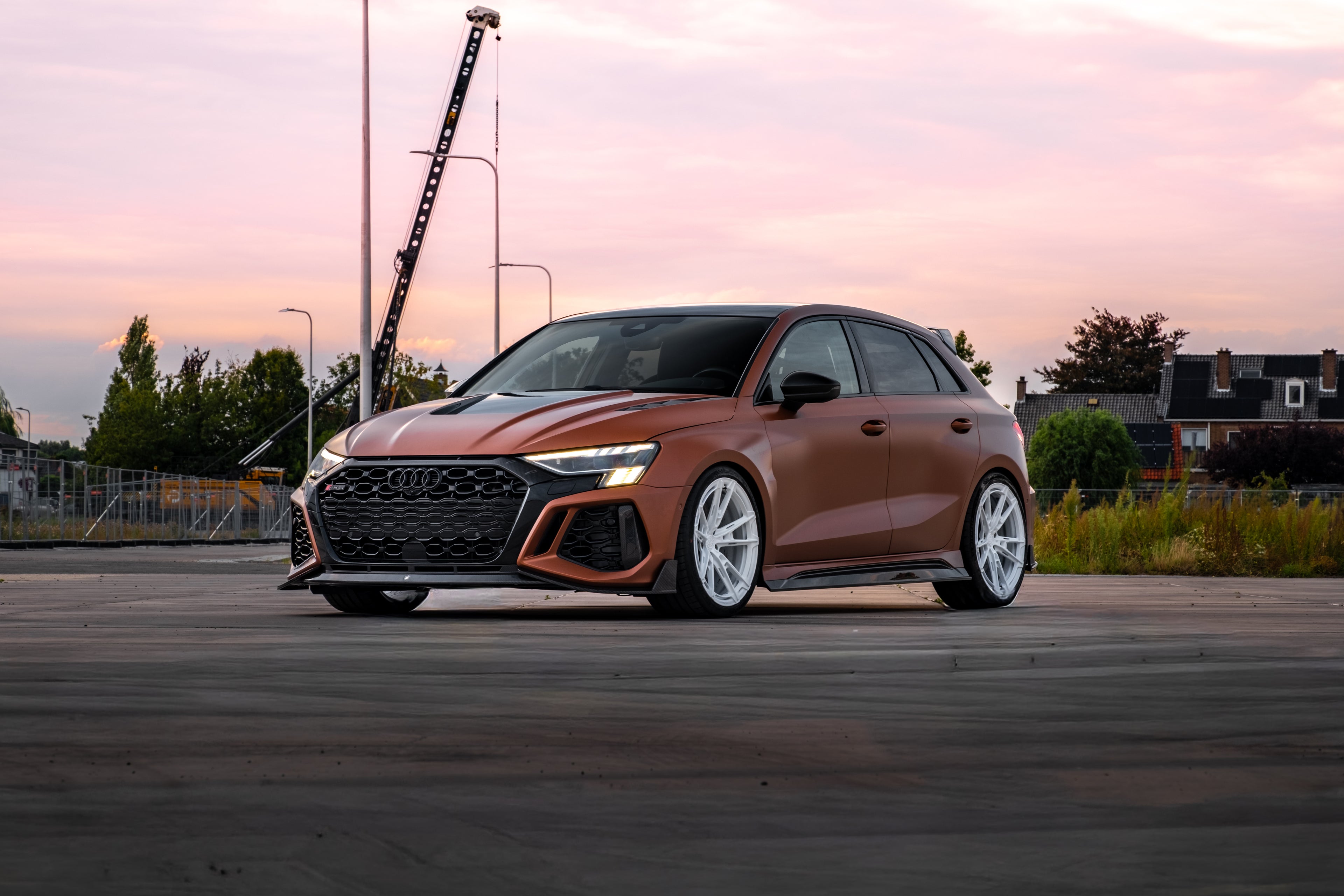 Bronze Audi RS3 Sportback featuring a REVOZPORT carbon fiber body kit with front lip and side skirts, paired with white multi-spoke forged wheels, parked at sunset in an open urban setting.