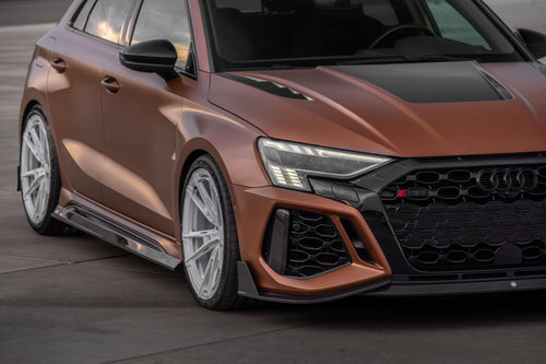 Bronze Audi RS3 Sportback featuring a REVOZPORT carbon fiber body kit with front lip and side skirts, paired with white multi-spoke forged wheels, parked at sunset in an open urban setting.