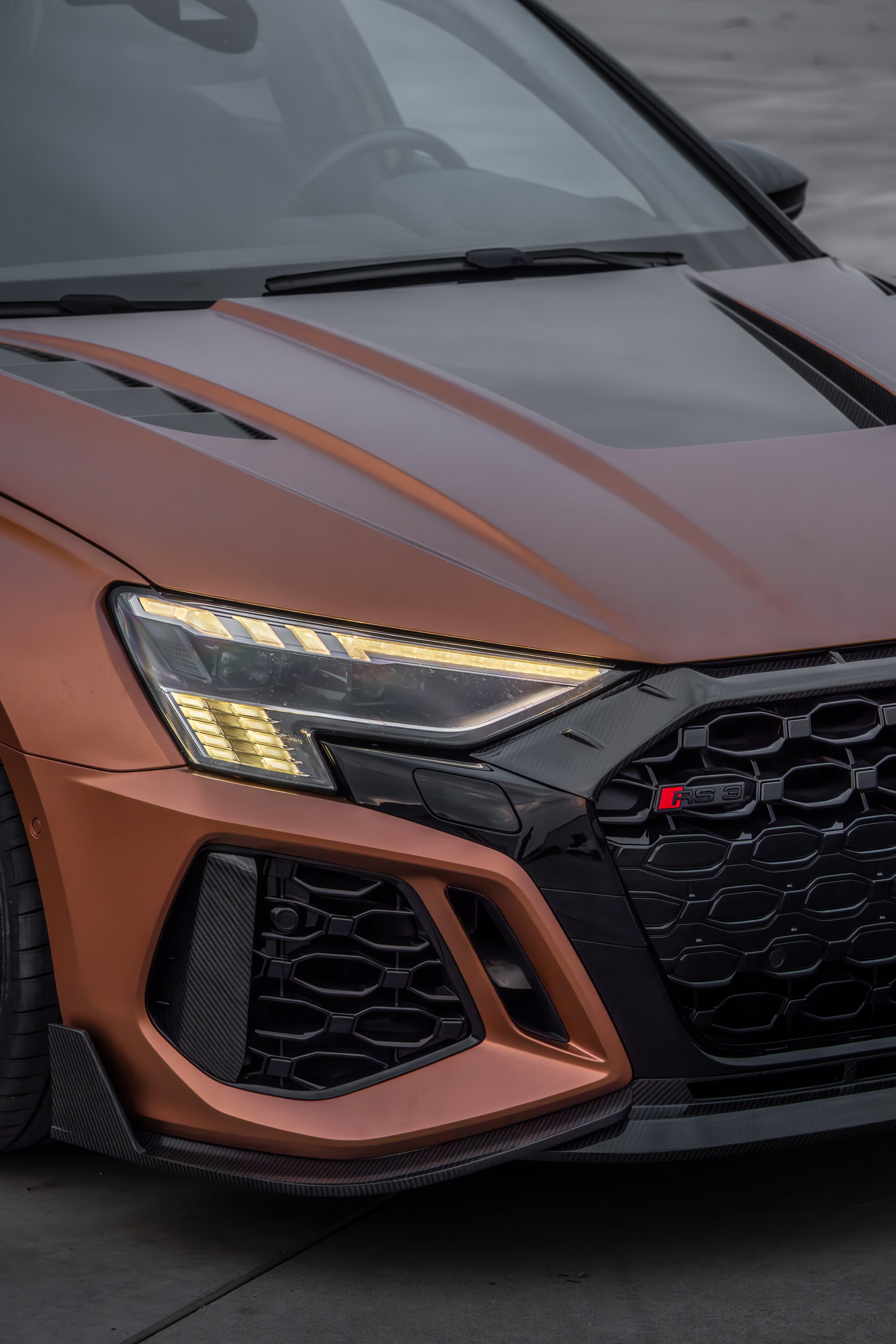 Bronze Audi RS3 Sportback featuring a REVOZPORT carbon fiber body kit with front lip and side skirts, paired with white multi-spoke forged wheels, parked at sunset in an open urban setting.