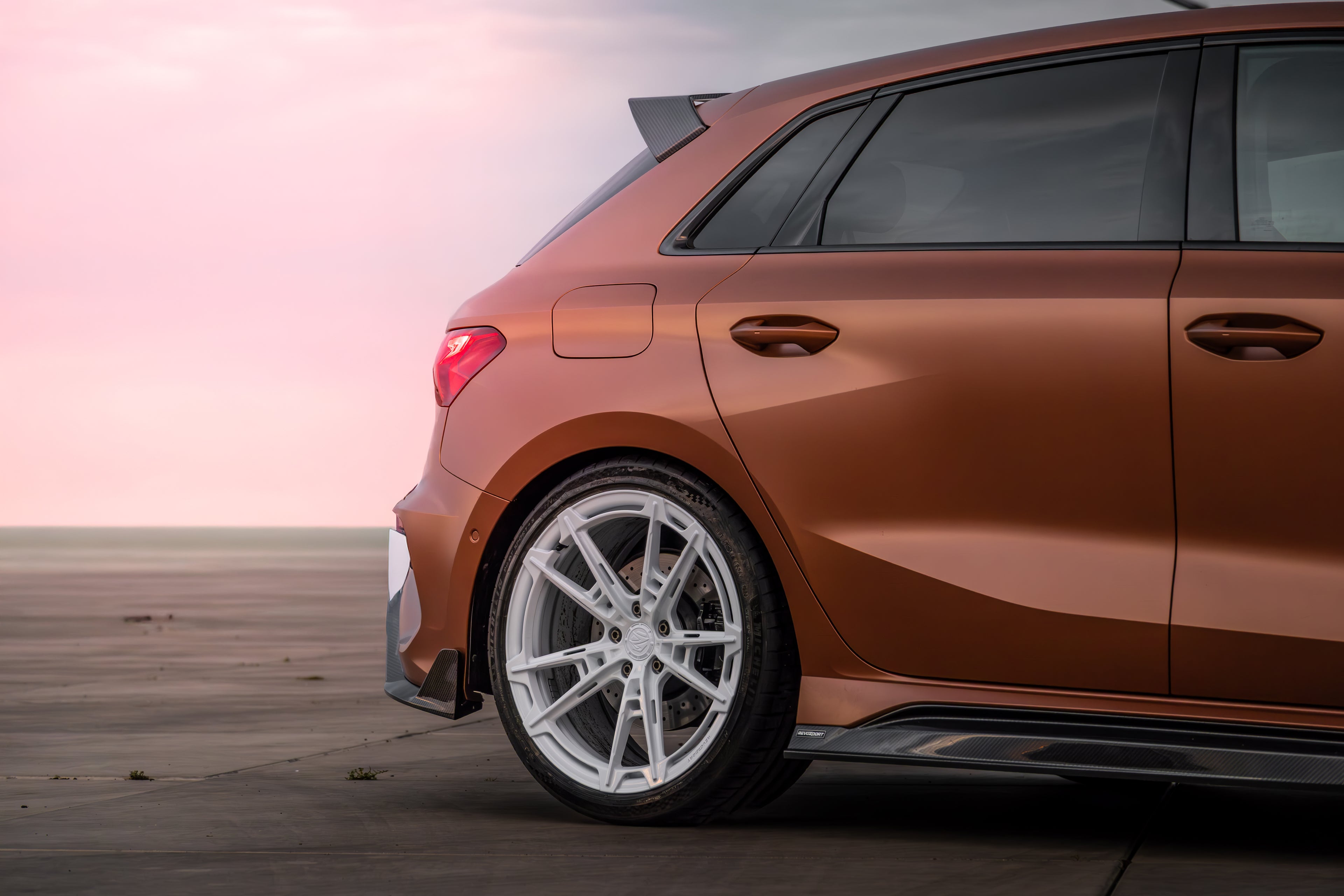 Bronze Audi RS3 Sportback featuring a REVOZPORT carbon fiber body kit with front lip and side skirts, paired with white multi-spoke forged wheels, parked at sunset in an open urban setting.