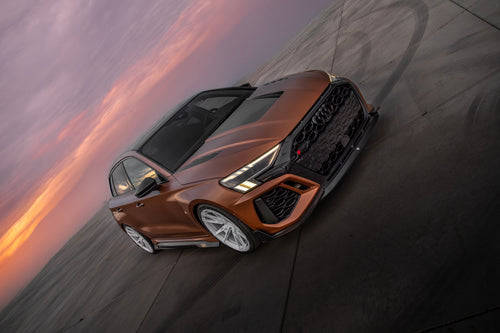 Bronze Audi RS3 Sportback featuring a REVOZPORT carbon fiber body kit with front lip and side skirts, paired with white multi-spoke forged wheels, parked at sunset in an open urban setting.