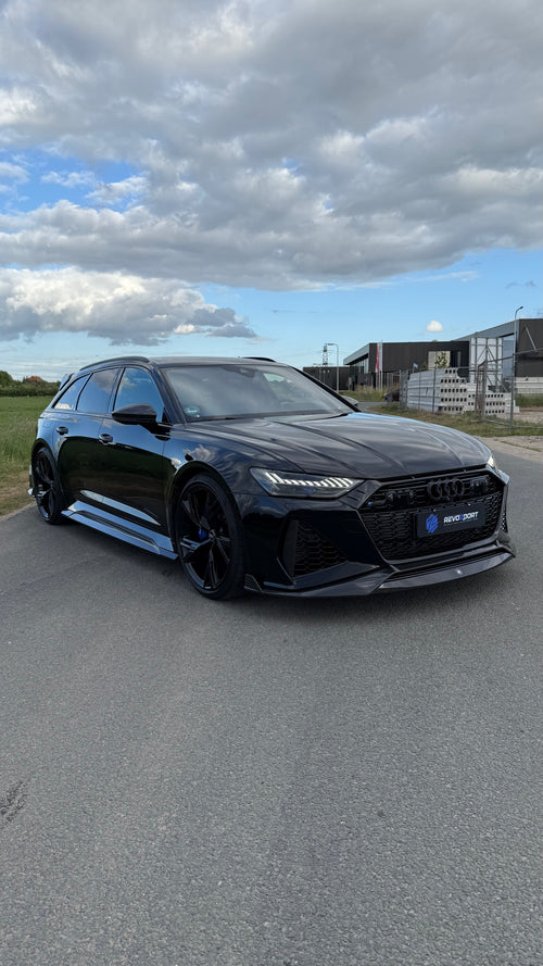 Black Audi RS6 Avant featuring a REVOZPORT carbon fiber body kit with front lip, side skirts, aggressive aero styling, and blacked-out wheels parked on an open road.