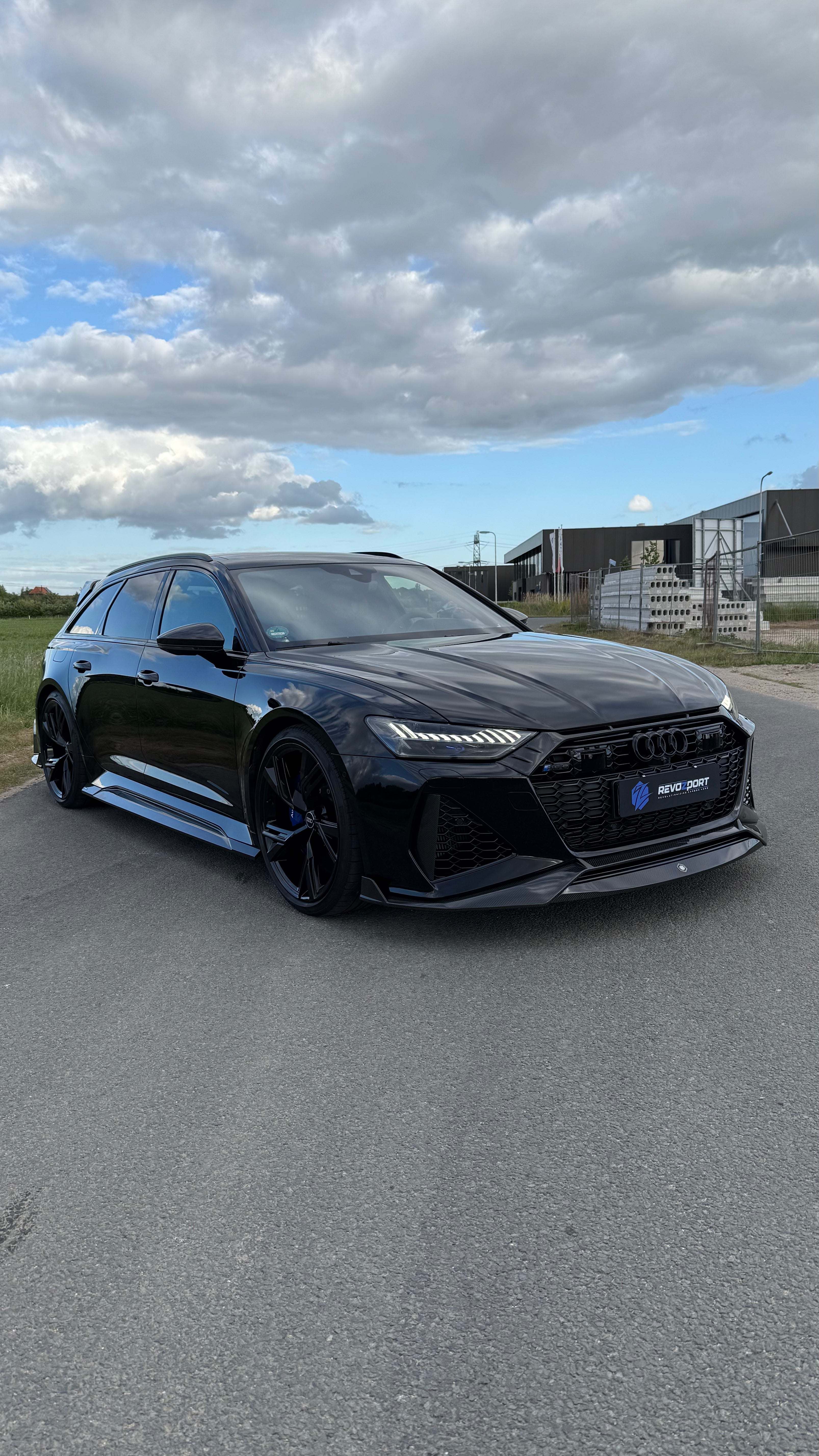 Black Audi RS6 Avant featuring a REVOZPORT carbon fiber body kit with front lip, side skirts, aggressive aero styling, and blacked-out wheels parked on an open road.