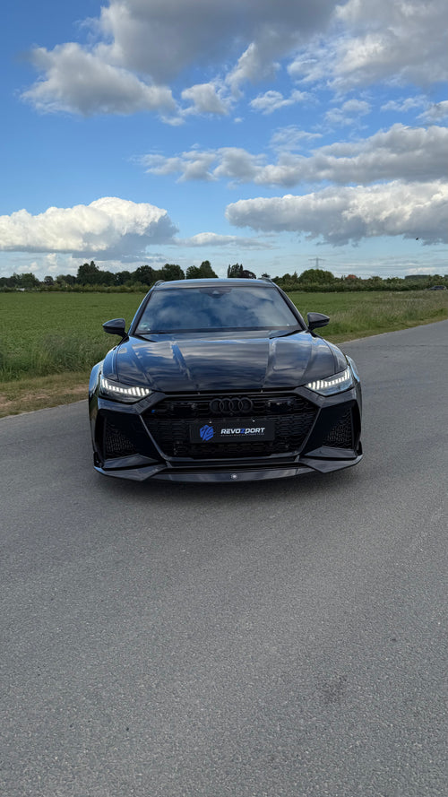 Black Audi RS6 Avant featuring a REVOZPORT carbon fiber body kit with front lip, side skirts, aggressive aero styling, and blacked-out wheels parked on an open road.
