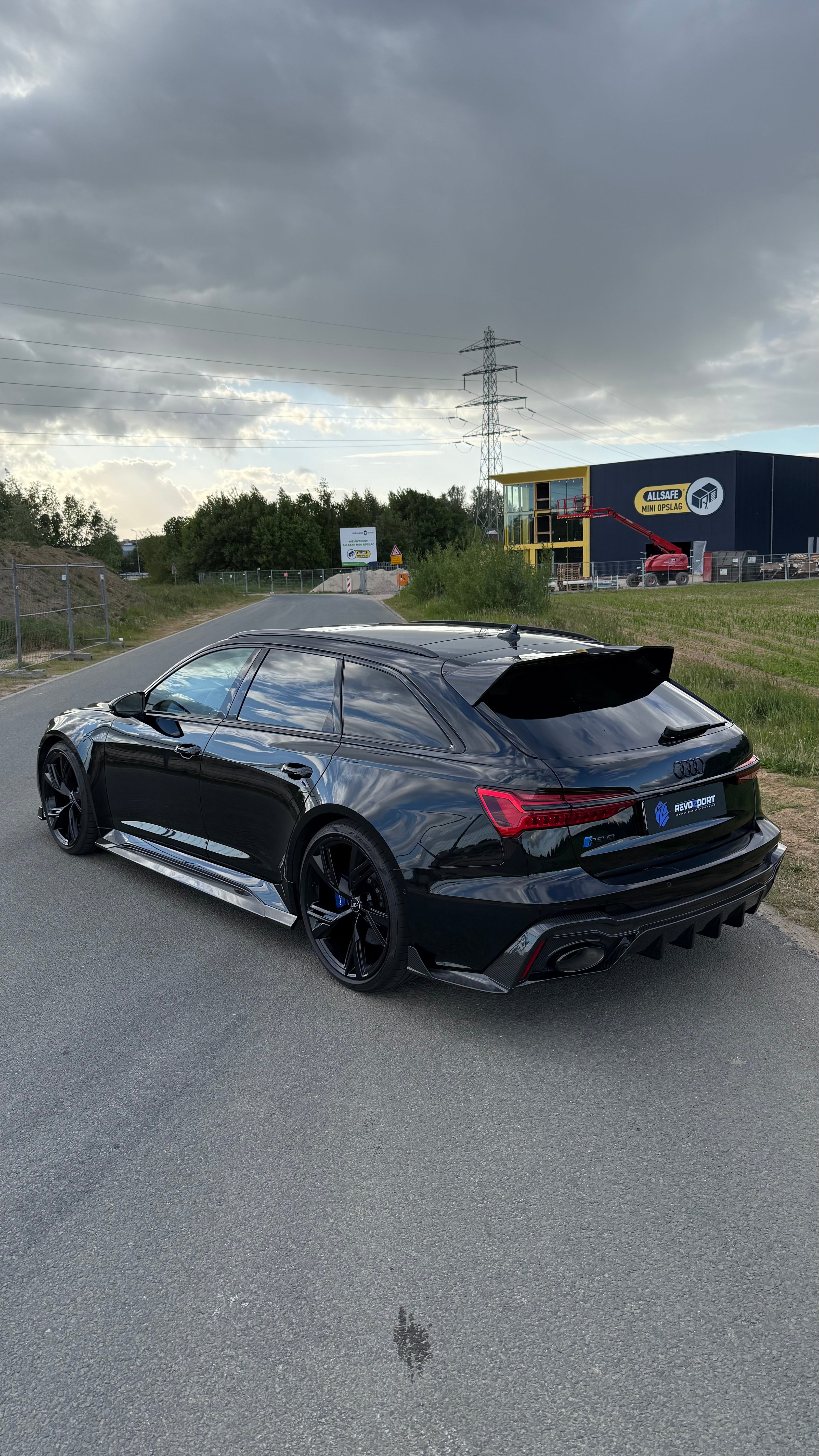 Black Audi RS6 Avant featuring a REVOZPORT carbon fiber body kit with front lip, side skirts, aggressive aero styling, and blacked-out wheels parked on an open road.