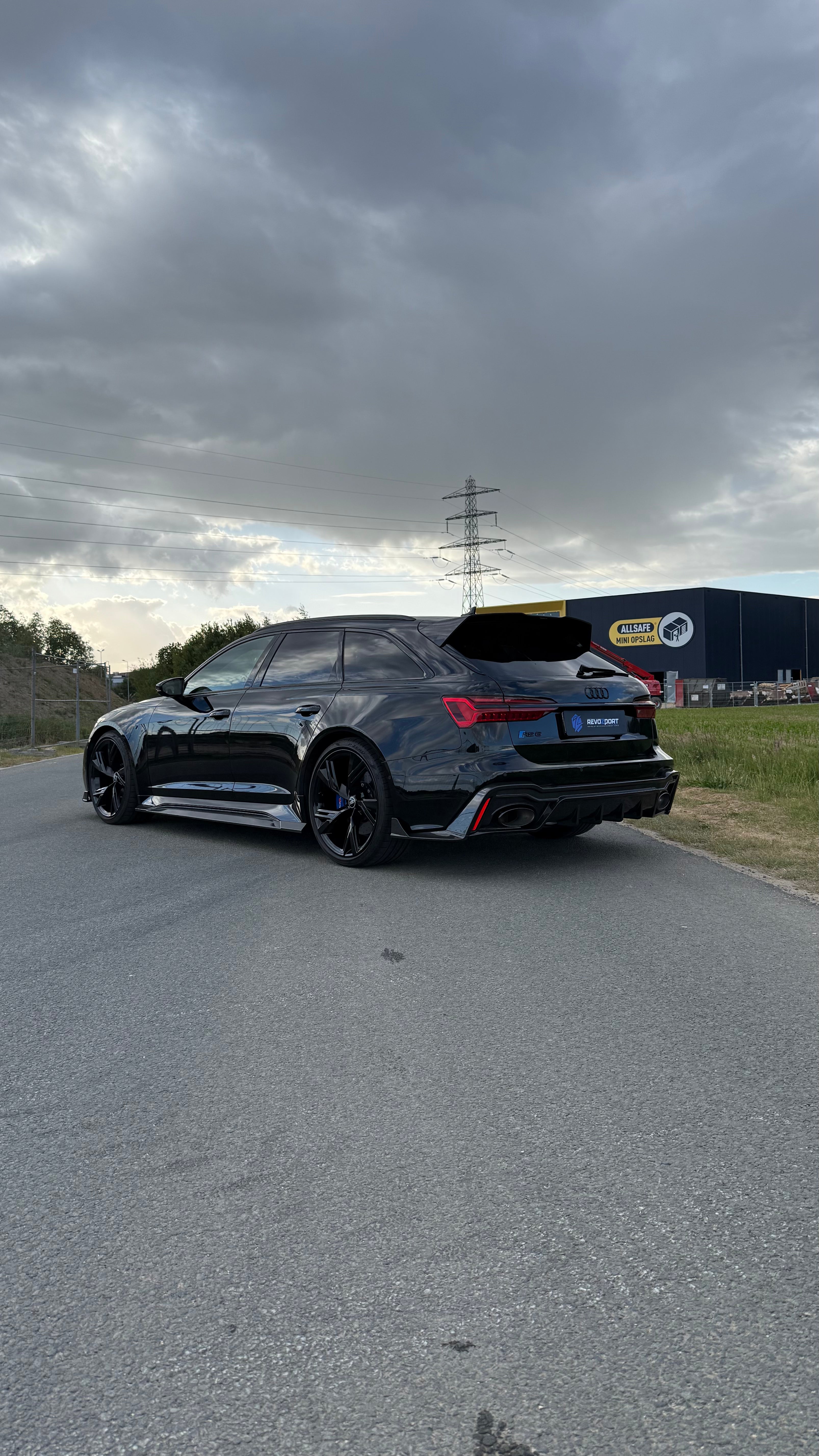 Black Audi RS6 Avant featuring a REVOZPORT carbon fiber body kit with front lip, side skirts, aggressive aero styling, and blacked-out wheels parked on an open road.